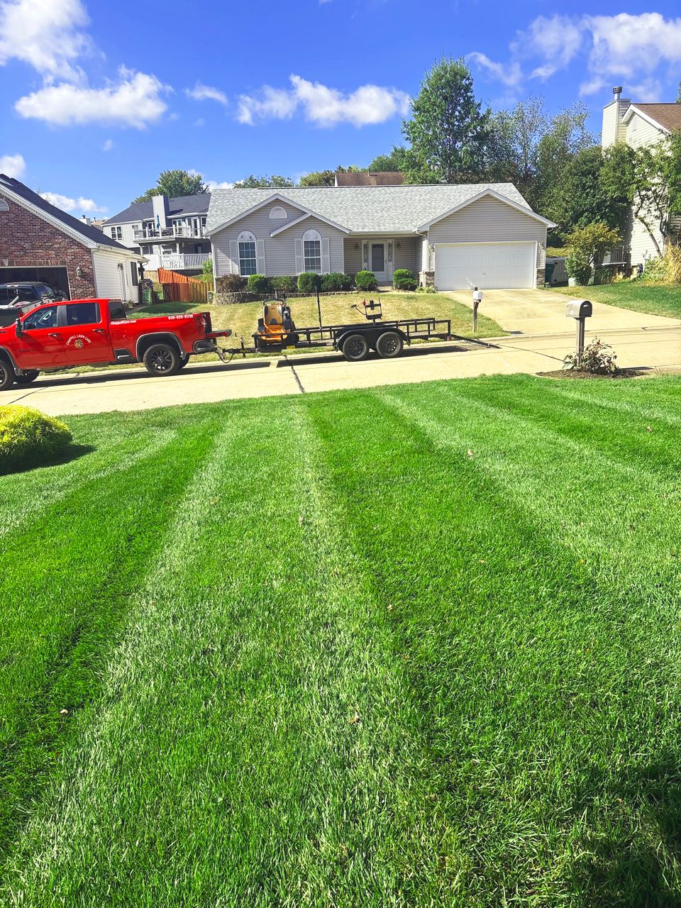 Green striped lawn in front of a light gray house. Red truck and trailer parked on the driveway.