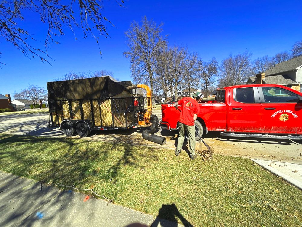 Lawn care worker loading debris into a trailer next to a red truck on a residential street.