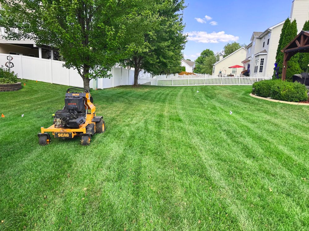 Yellow lawnmower on lush green grass, mowing a backyard lawn with a white fence.