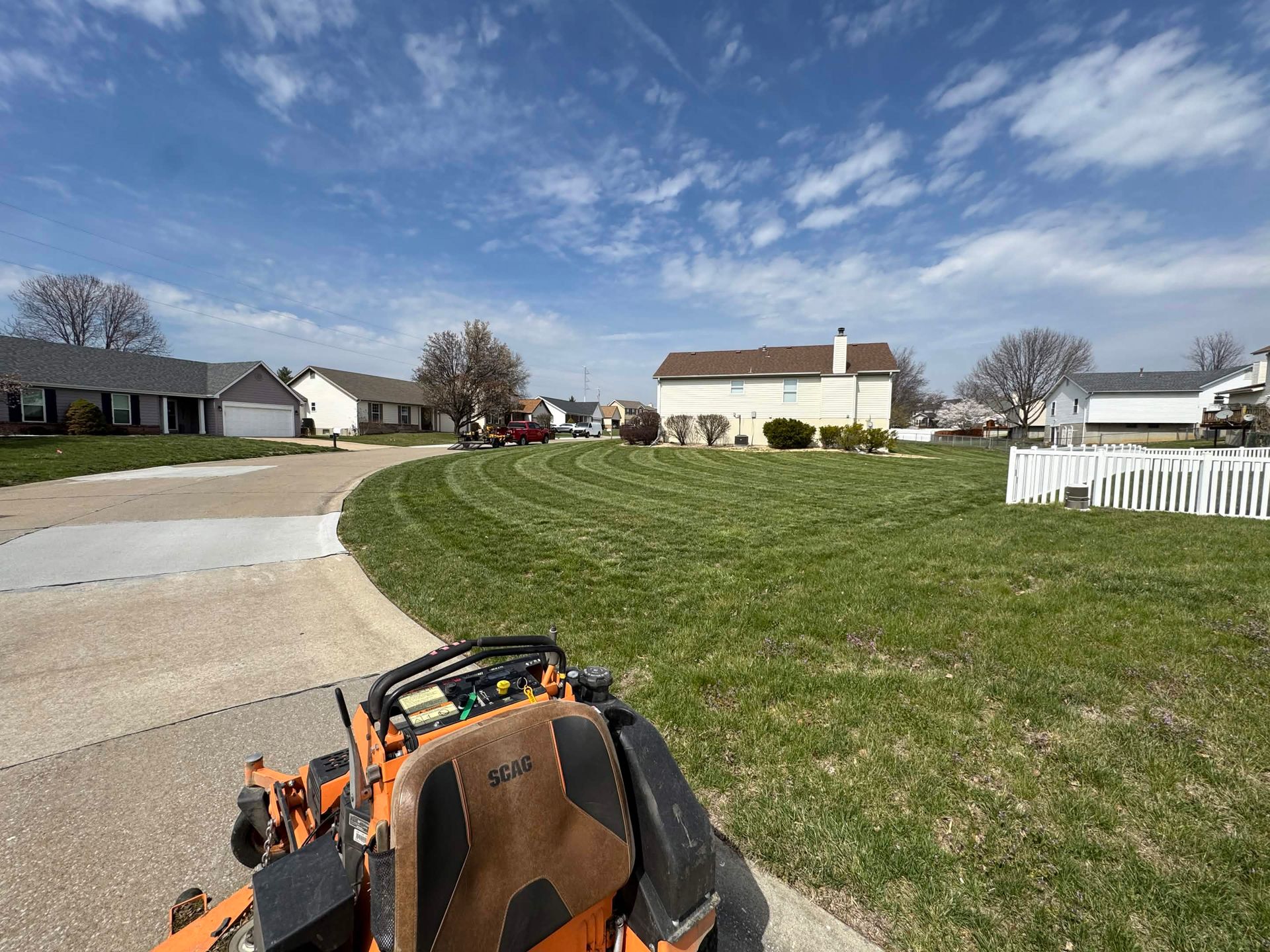 Front yard with mowed grass, curved driveway, white houses, and a landscaping machine in the foreground