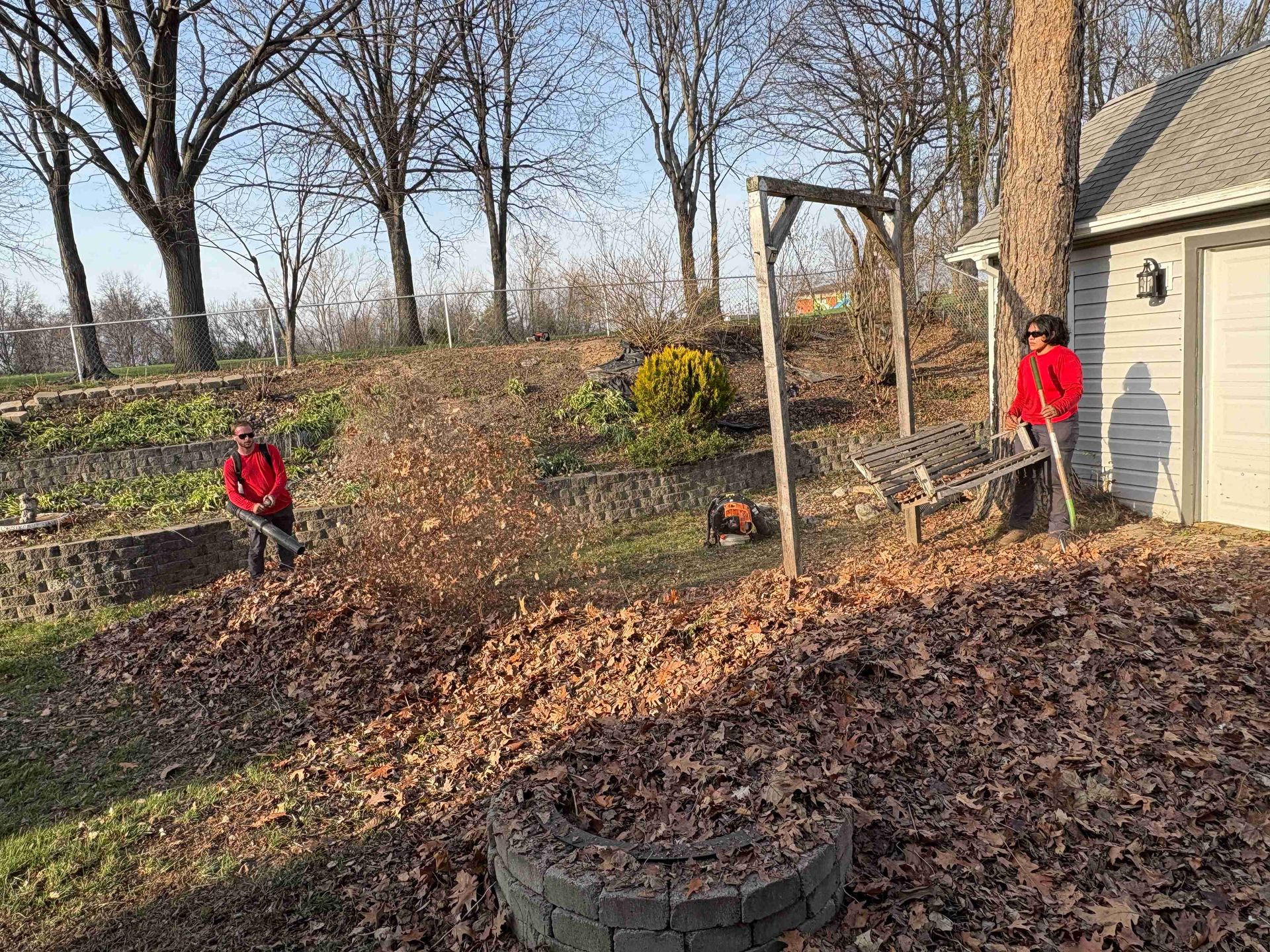 Two people in red jackets outdoors near a house, with fallen leaves and bare trees in a yard.