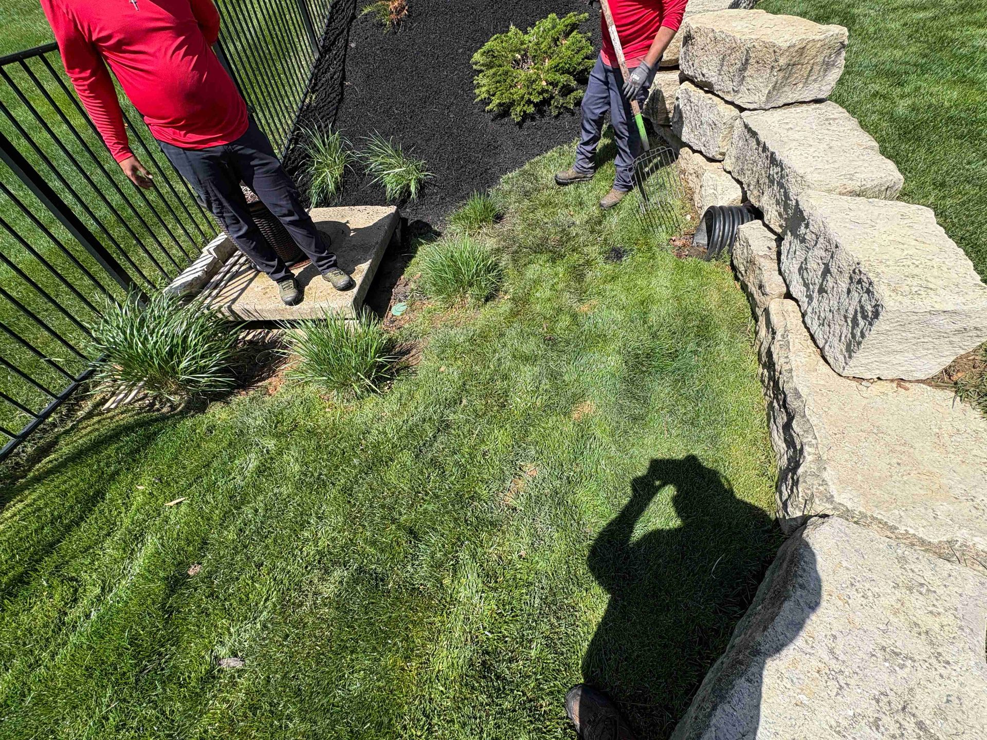 Two workers in red shirts standing by a black fence and stone wall on a grassy slope.