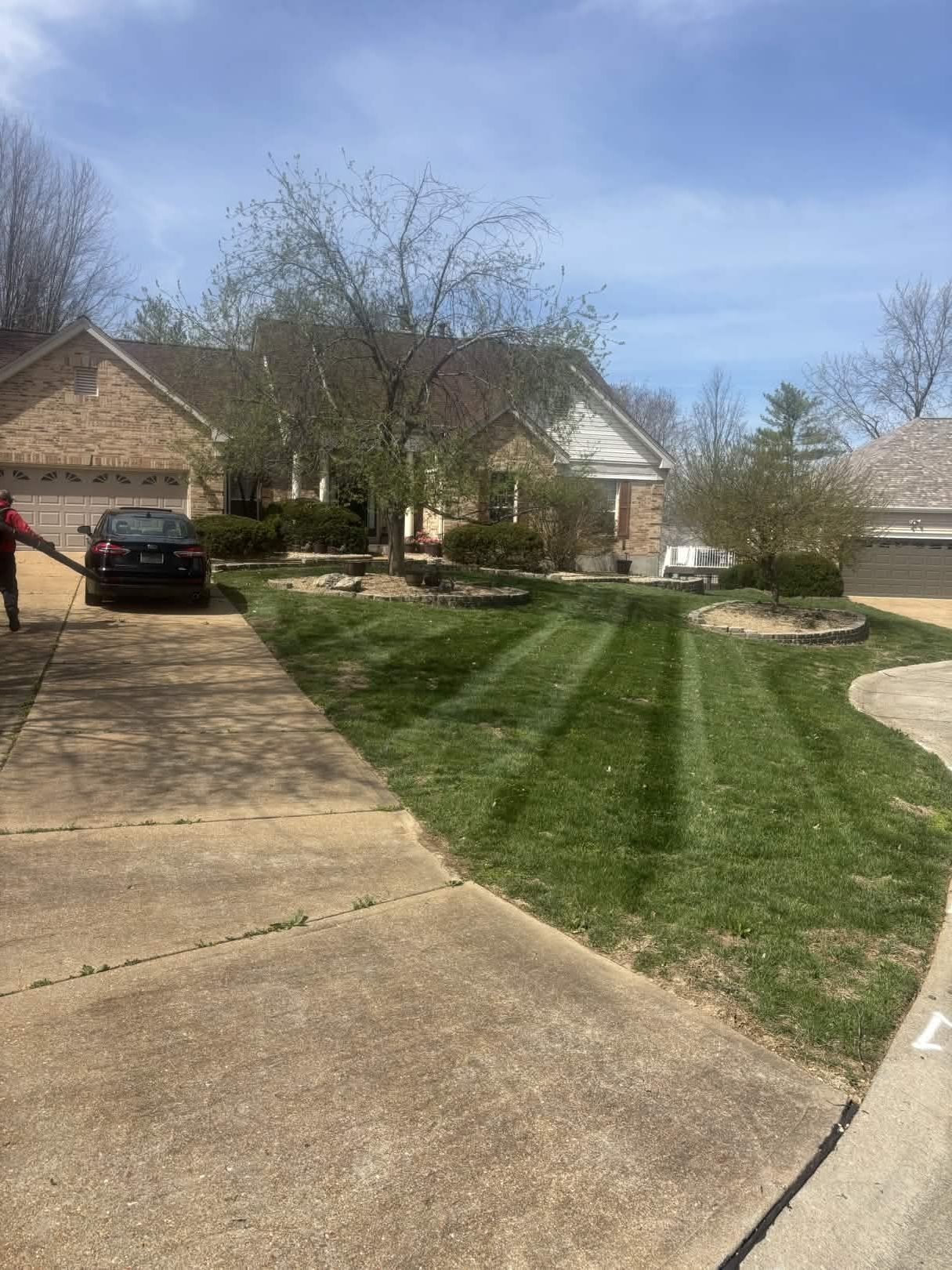 Suburban driveway and lawn with a parked car, houses, and trees on a clear day