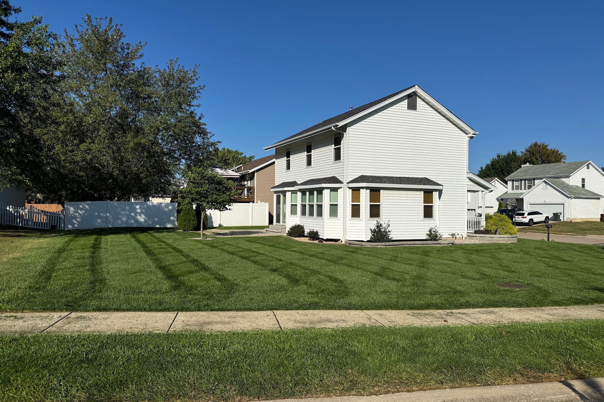 White two-story house with green lawn, cut in striped pattern, under a blue sky.