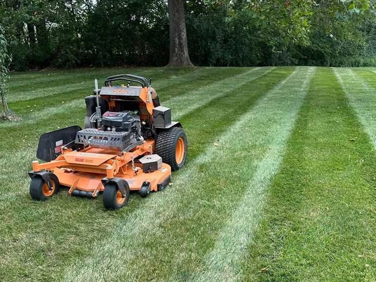 Orange riding lawnmower mowing a lawn, creating striped patterns. Green grass, trees in the background.
