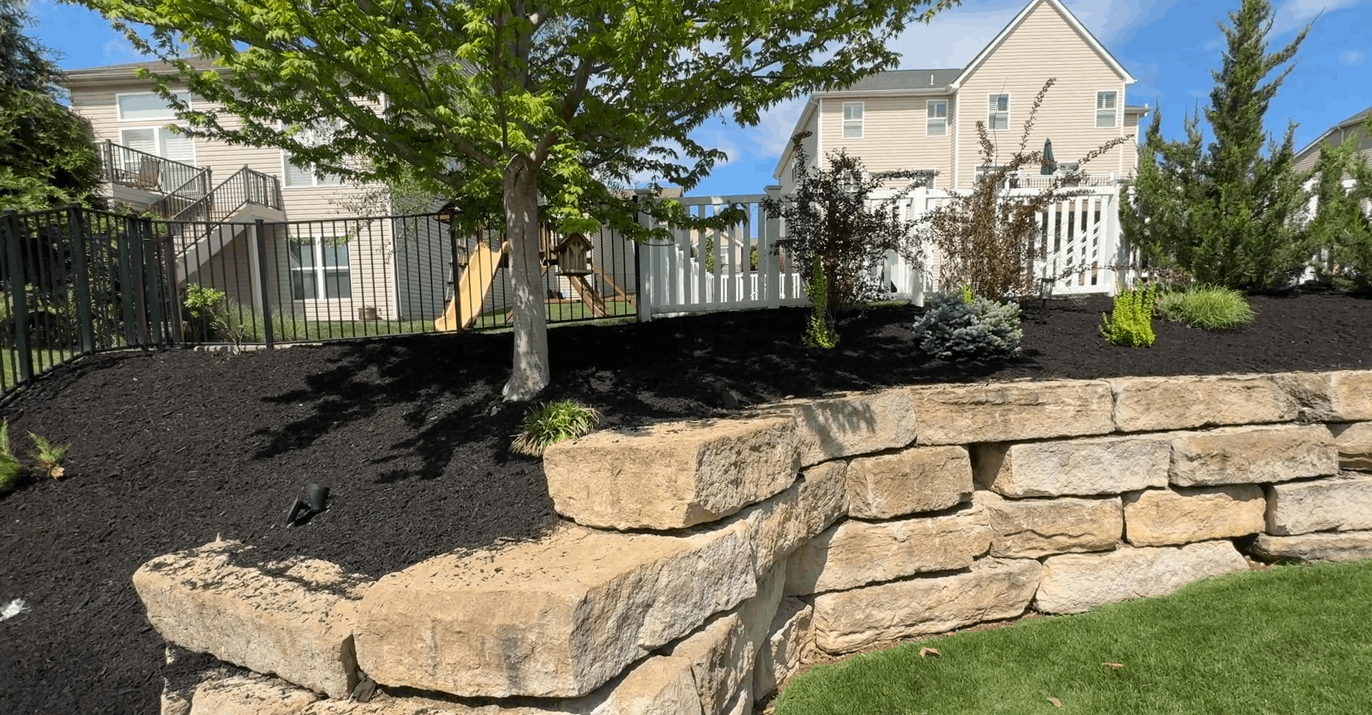 Backyard with a stone retaining wall, mulched bed, tree, and white house in the background.