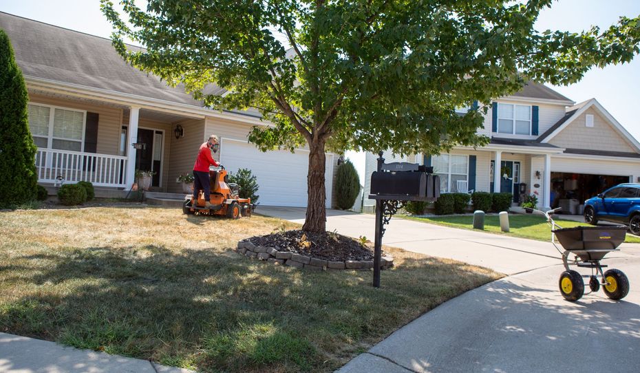 Orange riding lawnmower mowing a lawn, creating striped patterns. Green grass, trees in the background.