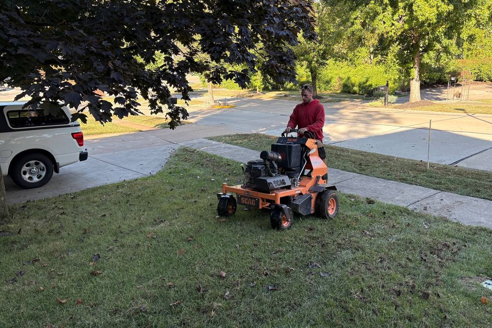 Person operating a zero-turn lawnmower on a lawn next to a driveway; truck parked nearby.