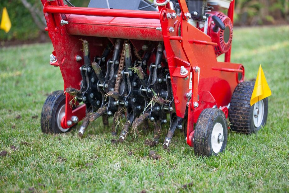 Red tractor-mounted aerator with rotating tines on grass, turning soil in a lawn.