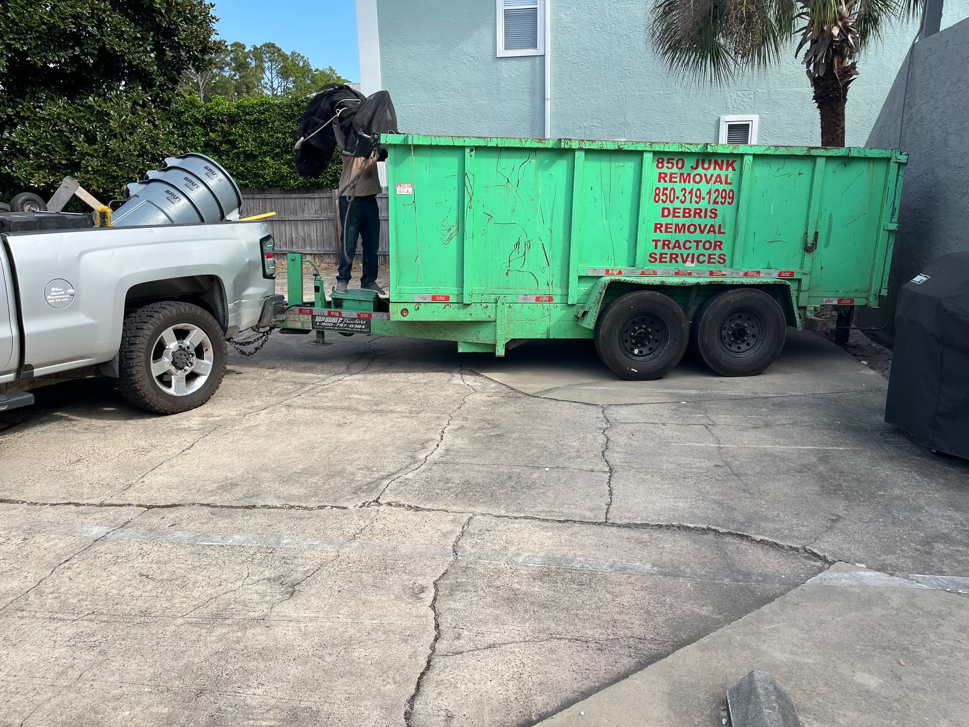 A green dumpster is being towed by a truck in a driveway.