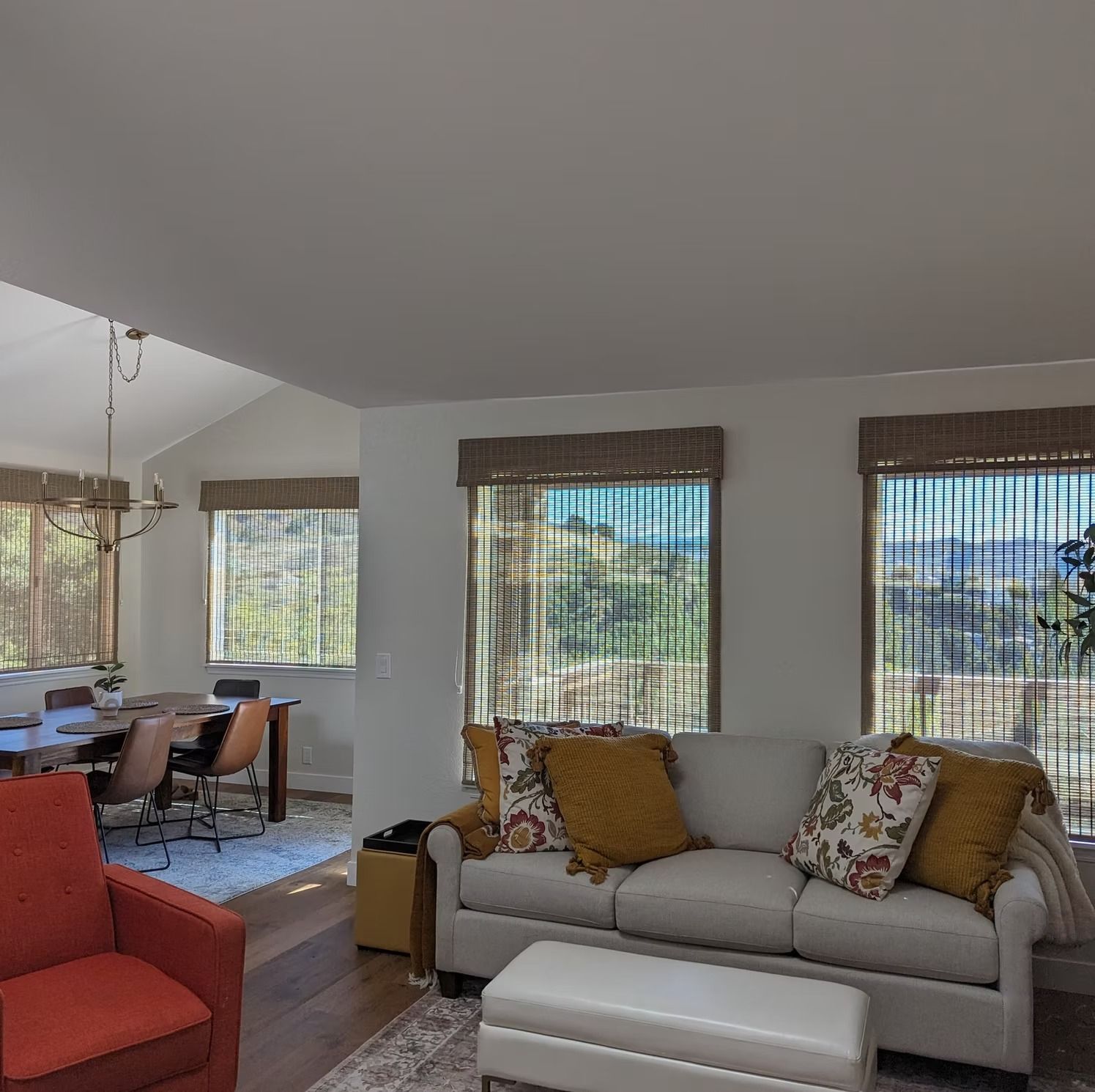 Bright living room with cream sofa, red chair, dining area, and large windows with woven blinds.