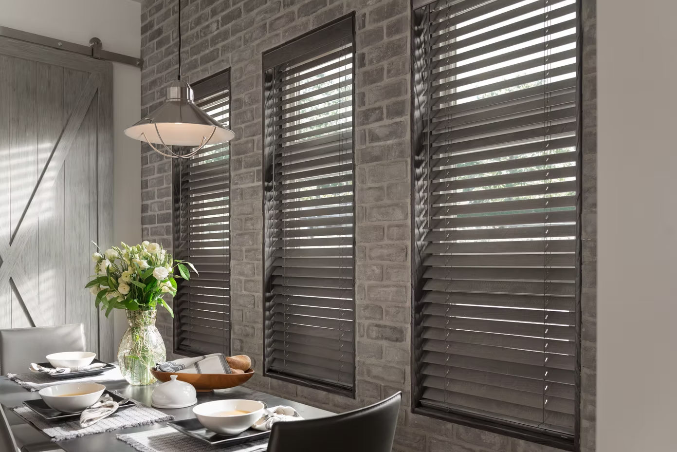 Dining table beside tall windows with dark blinds, white tableware, and a vase of flowers