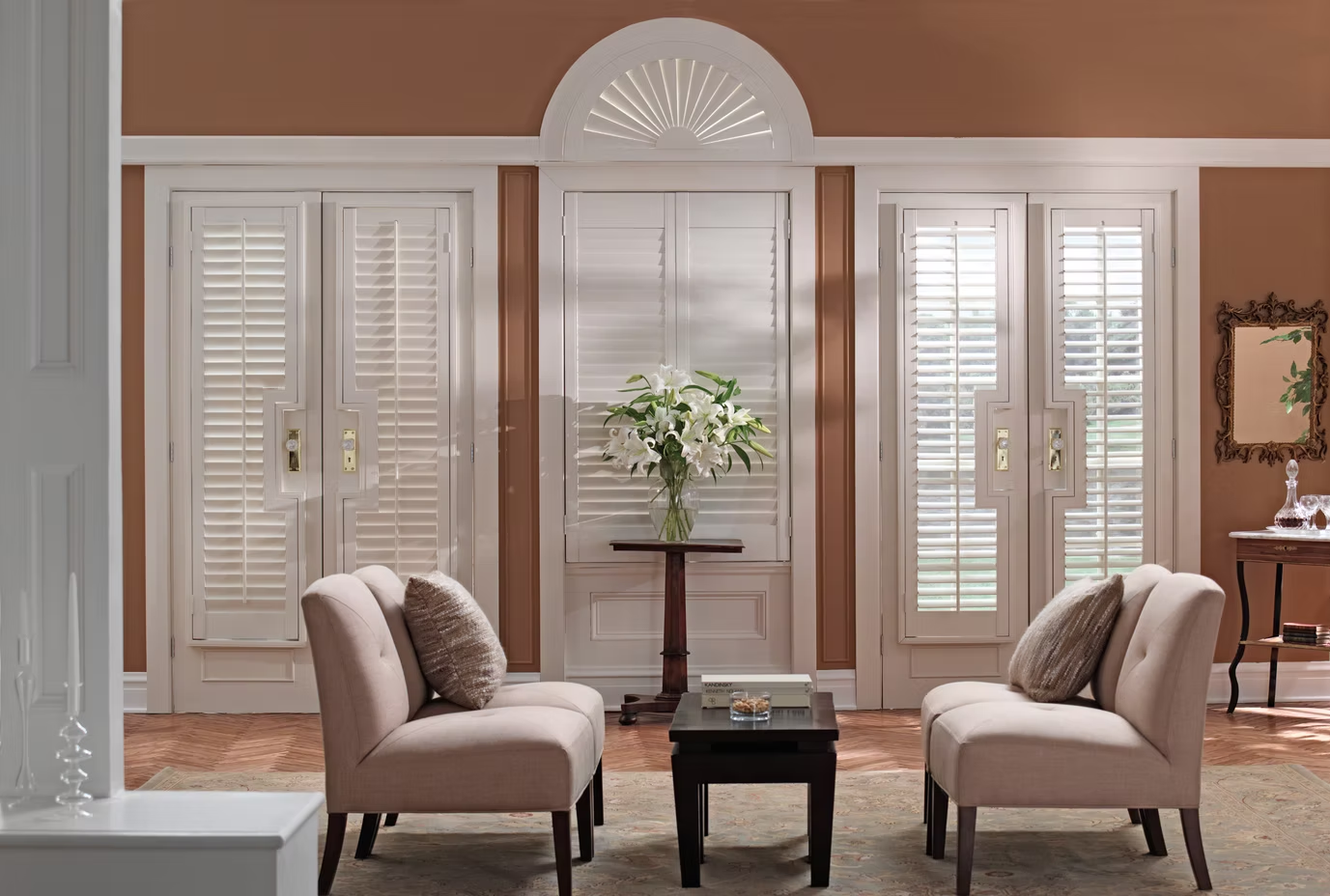 Elegant living room with two beige chairs, a small table, and bright shutters around a floral arrangement.