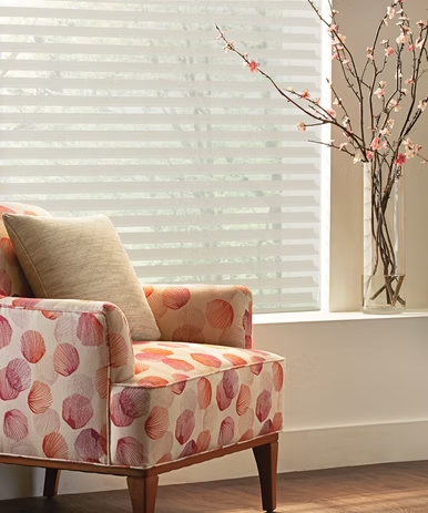 Patterned armchair by a bright window with white blinds and a vase of pink branches