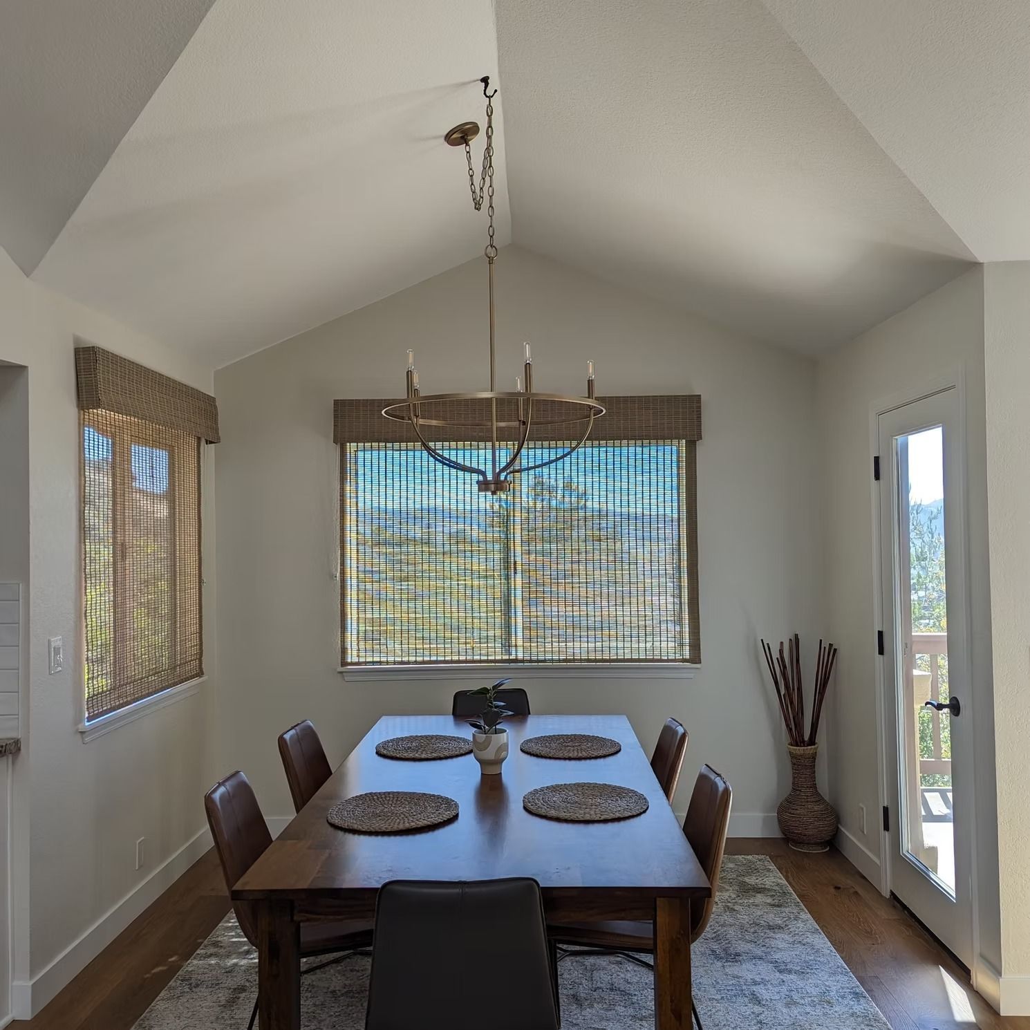 Dining room with wooden table, six chairs, and large windows with blinds and a hanging chandelier.