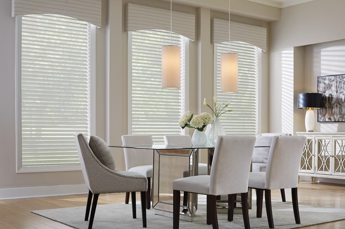 Bright dining room with table, upholstered chairs, white blinds, and a vase of flowers.