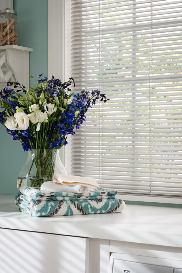 White table with blue and white flowers in a glass vase beside folded napkins by a sunlit window with blinds