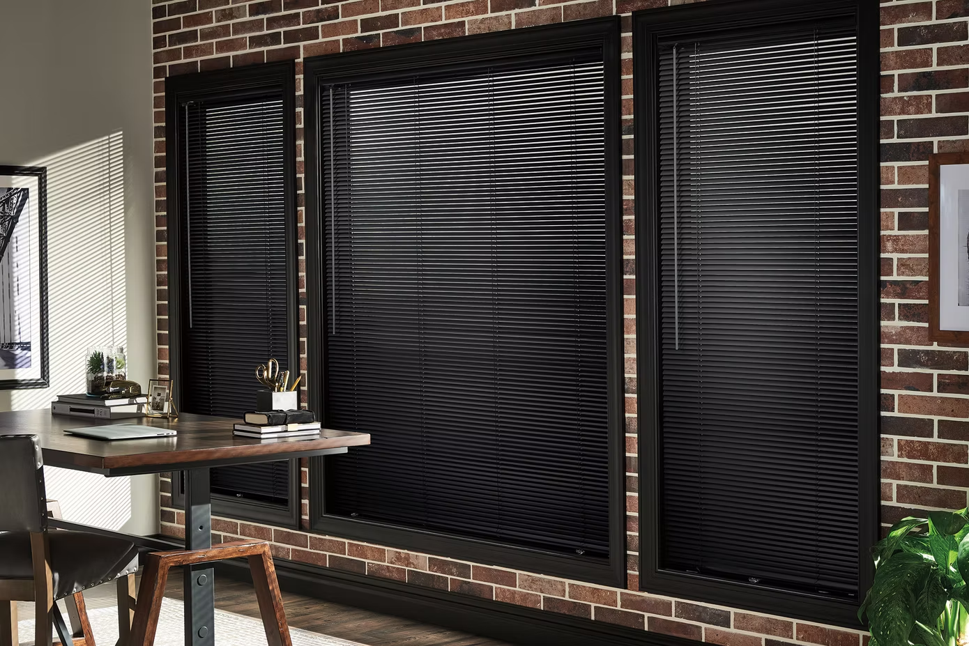 Brick-walled room with three large windows covered by black blinds and a small wooden desk near the left window