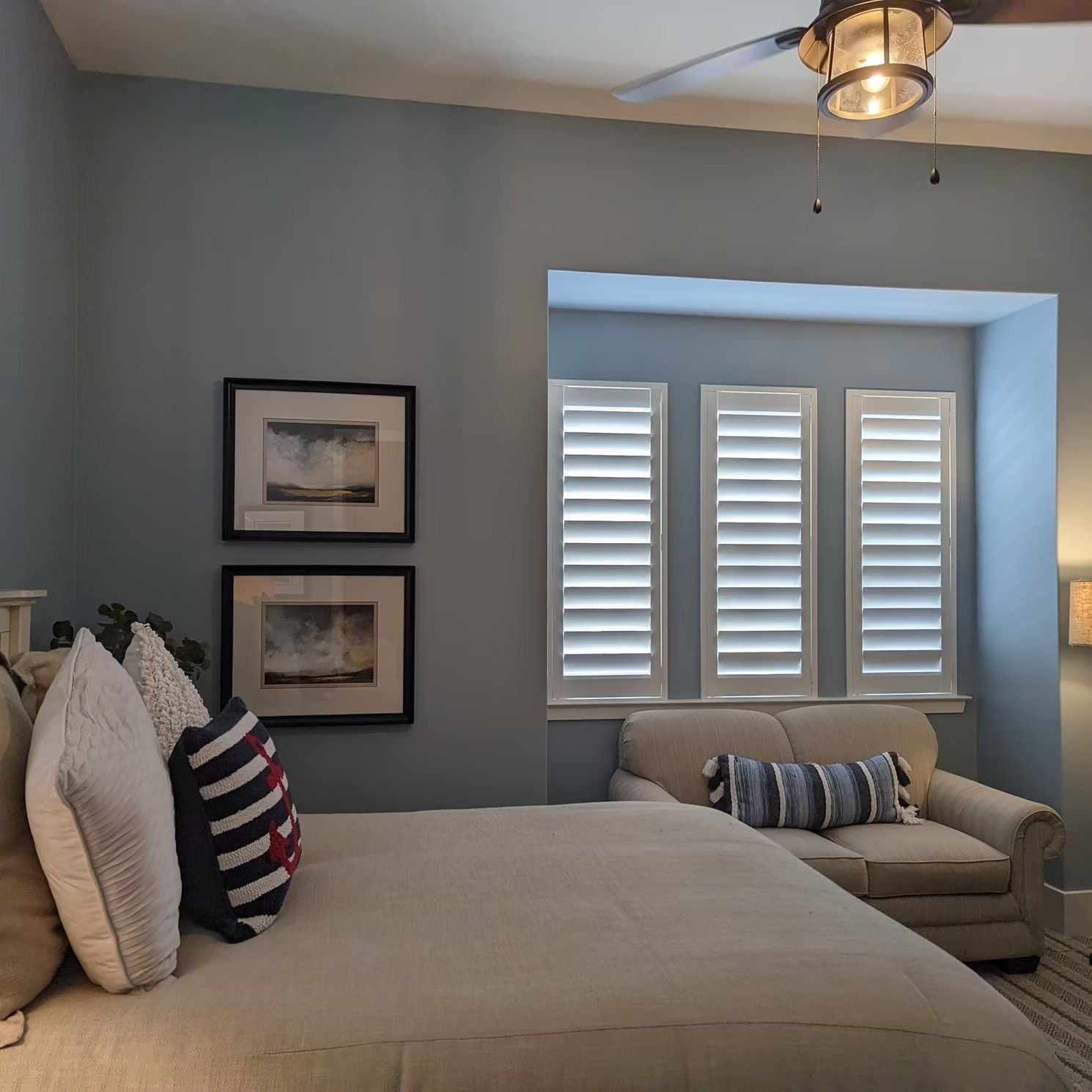 Neatly made bedroom with a large bed, light blue walls, shuttered window, and a small armchair by the window.
