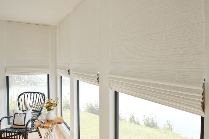 White pleated blinds covering tall windows in a bright room with a chair and small table