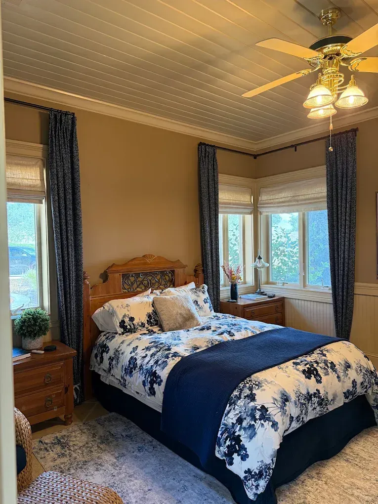 Cozy bedroom with blue floral bedspread, bay windows, dark curtains, and a ceiling fan with lights