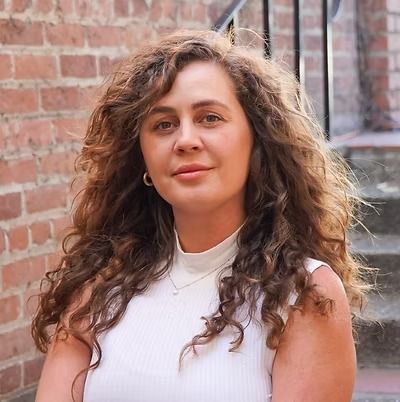 Portrait of a person with curly hair in a white sleeveless top, standing outdoors by a brick wall