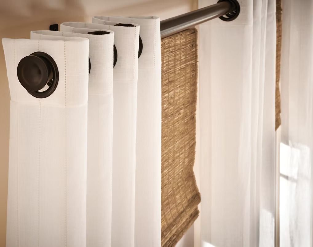 White curtains on a black curtain rod, with a woven tan shade behind them.