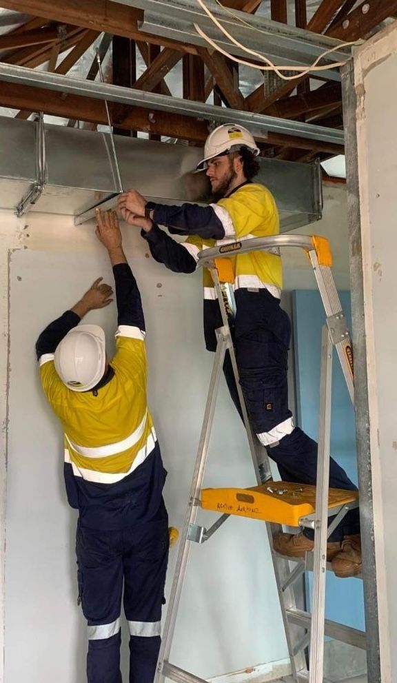 Two workers in yellow safety vests install a wall panel on a ladder in a construction site. — Active Airconz in Winnellie, NT