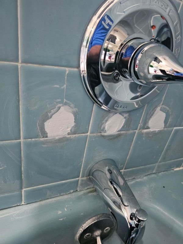 Blue bathroom tile wall with chipped patches around a chrome bathtub faucet and shower control