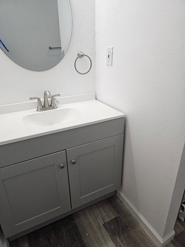 Gray bathroom vanity with sink, mirror, and towel ring against white walls and dark wood floor