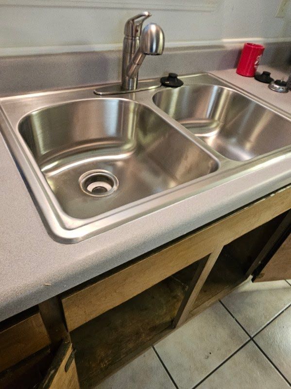 Stainless steel double-basin kitchen sink with faucet and beige tiled floor underneath.