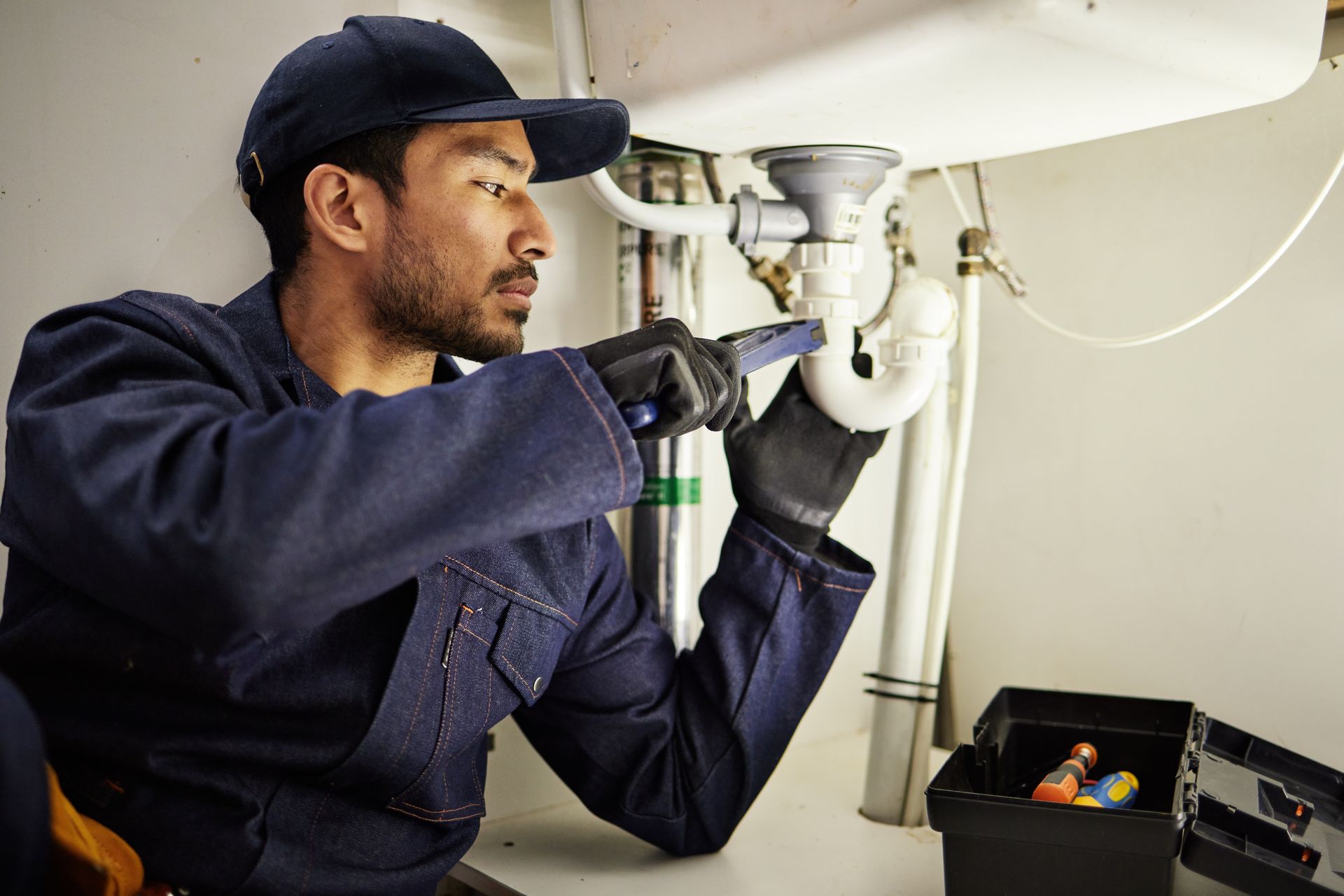 Plumber inspecting under-sink pipes with a flashlight and tools in a kitchen cabinet