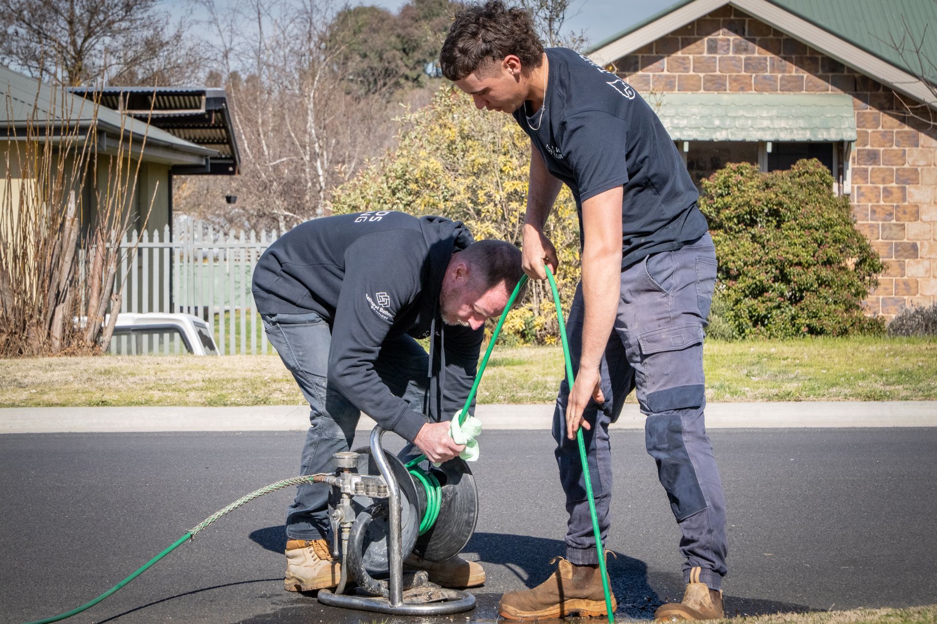 Two workers using a green hose and tool on a driveway in front of a house — T. Woods Plumbing in Bathurst, NSW