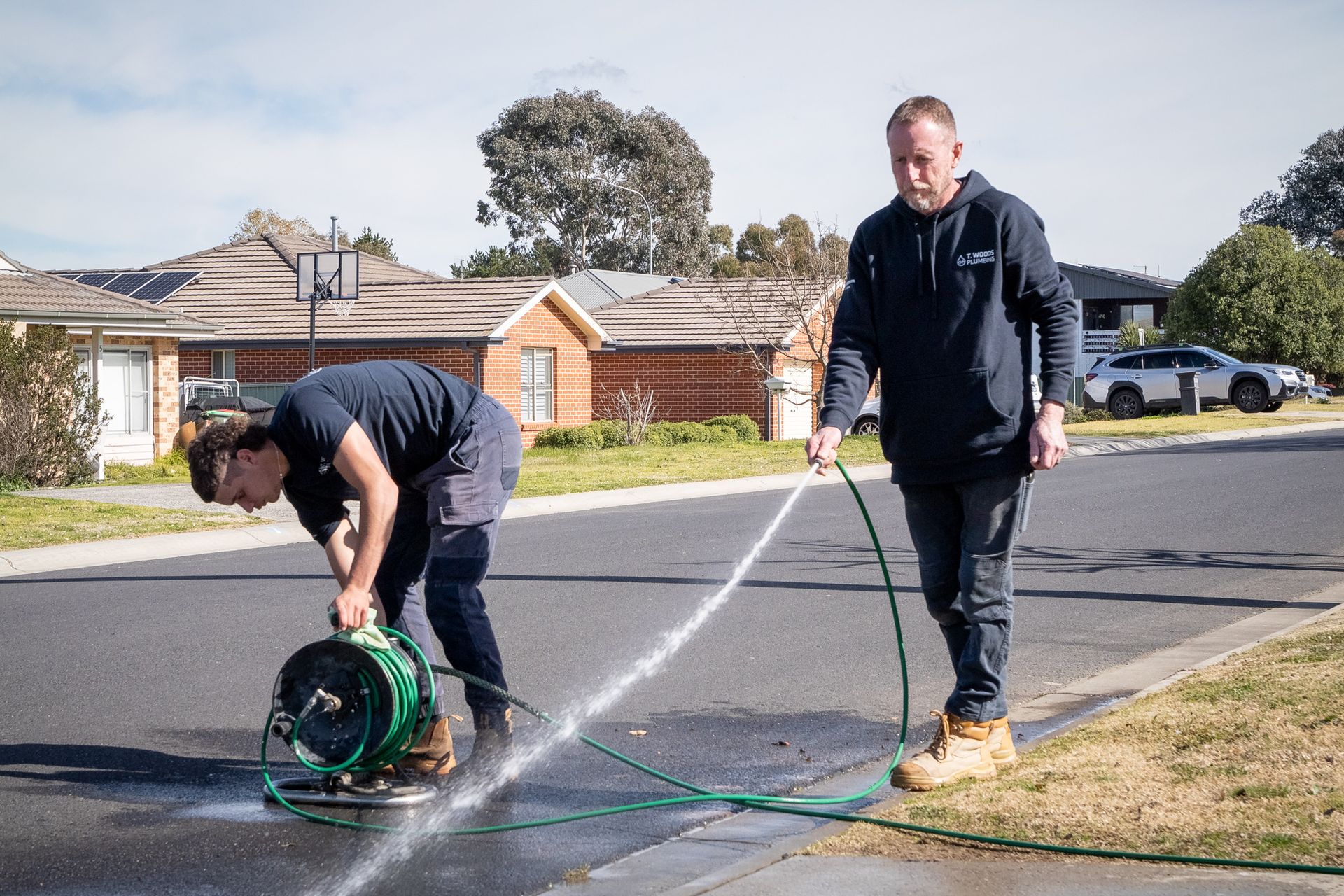 Two workers using a hose to clean a street with a hose reel on a suburban road. — T. Woods Plumbing in Bathurst, NSW
