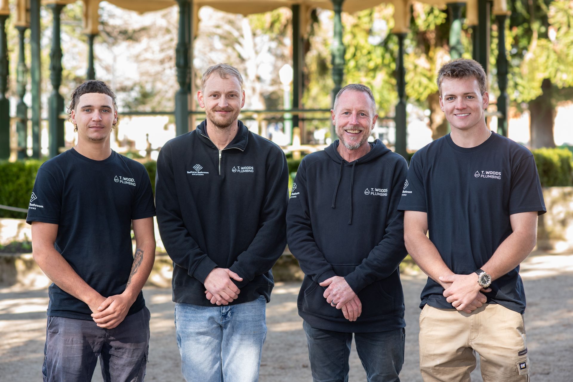 Four People Standing Outdoors in Black Shirts, Smiling — T. Woods Plumbing in Bathurst, NSW