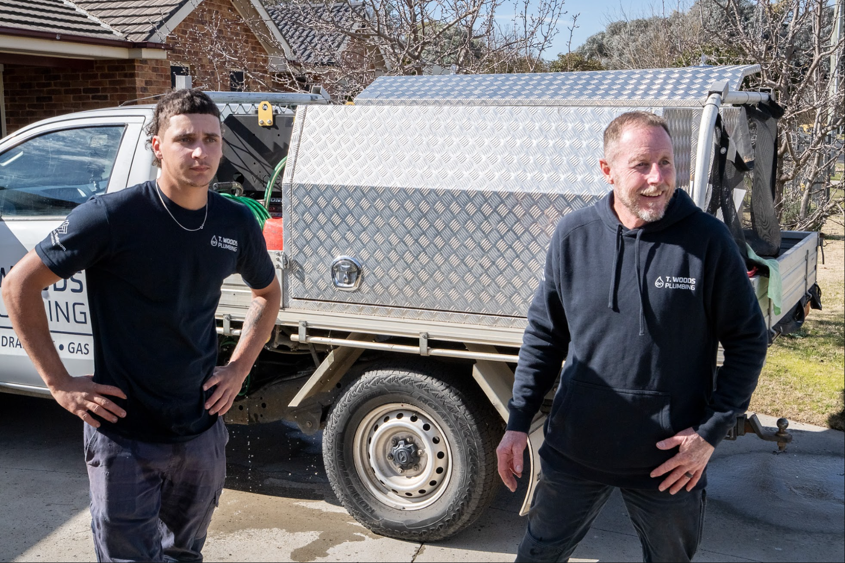 Two men stand beside a silver utility trailer in a driveway on a sunny day. — T. Woods Plumbing in Bathurst, NSW
