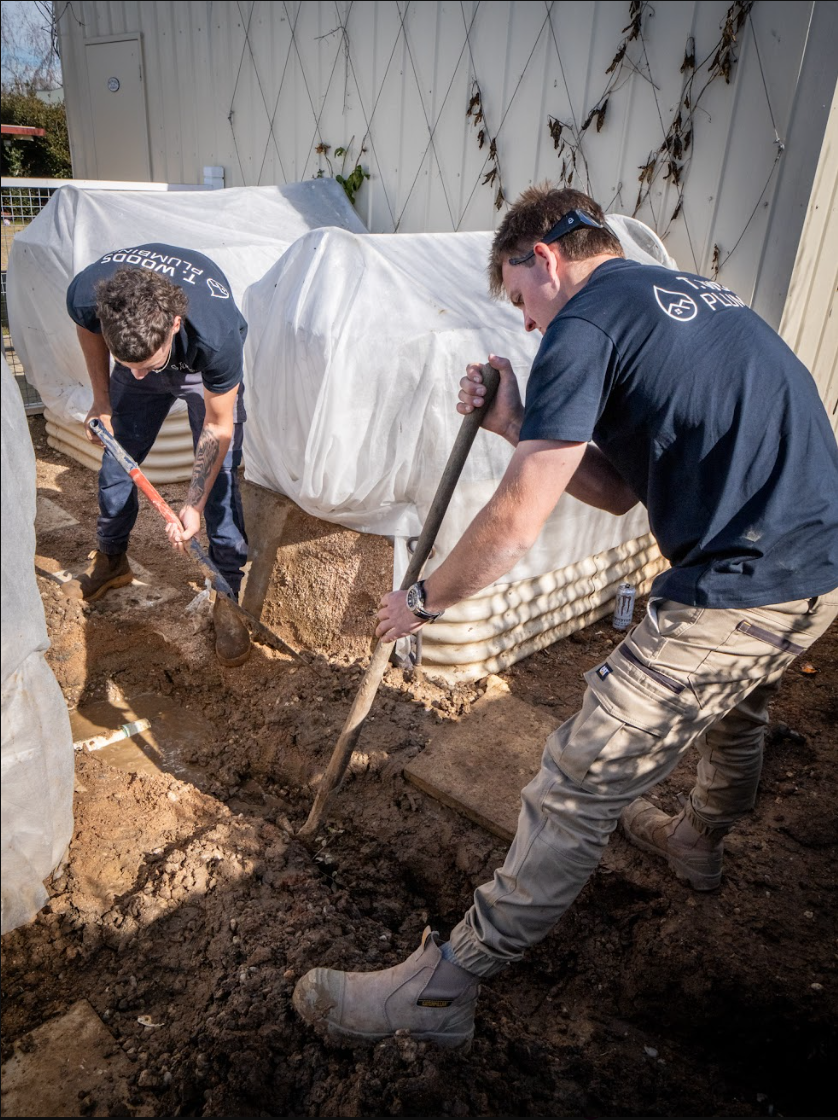 Two workers digging mud beside stacked sandbags and covered supplies near a wall — T. Woods Plumbing in Bathurst, NSW