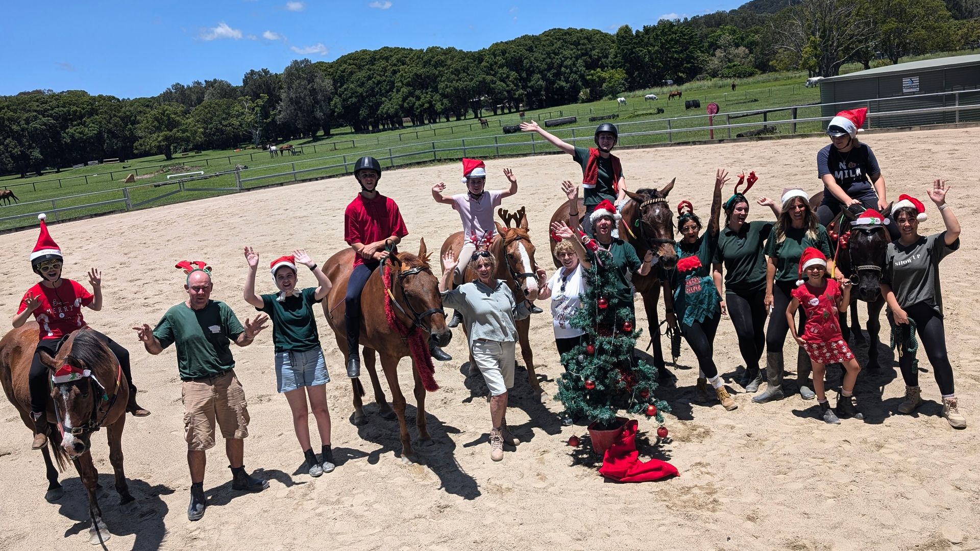 Group of people on and around horses, wearing Santa hats, waving, on a sandy field, with trees in the background.