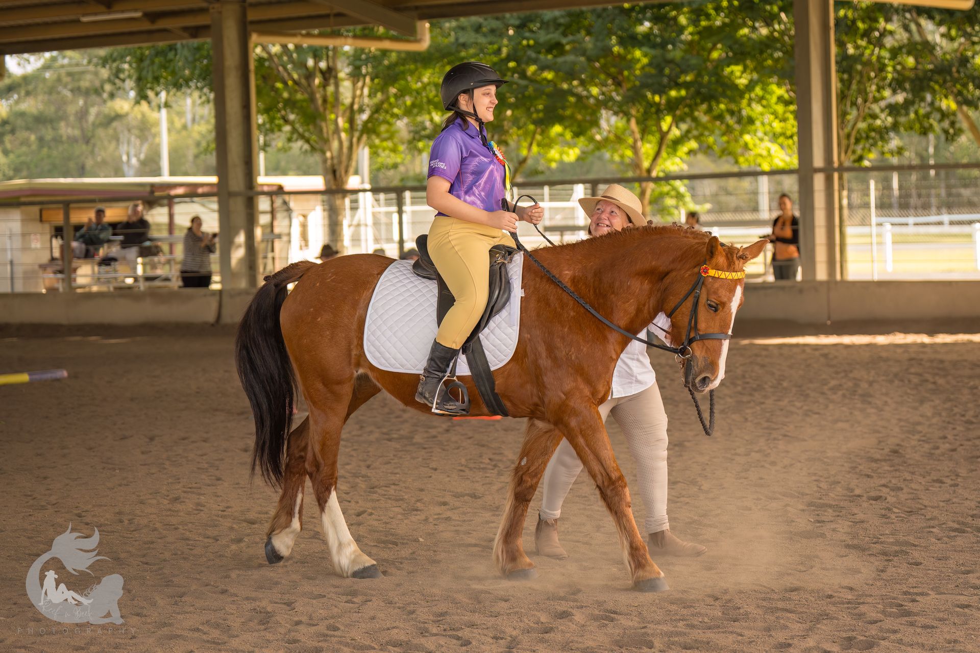 Woman riding chestnut horse in arena, being led by another person.