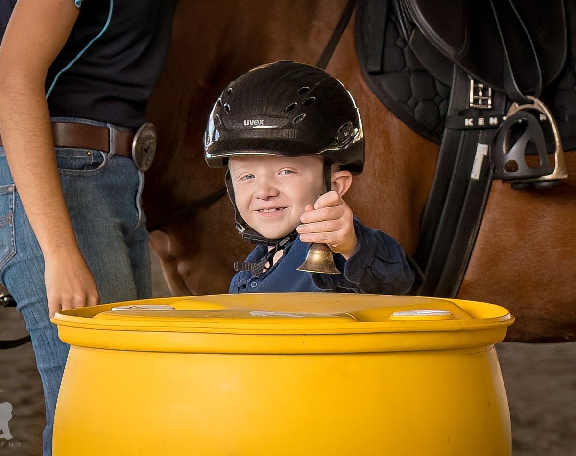 A child in a riding helmet holds a bell near a horse. A person in jeans stands beside them.