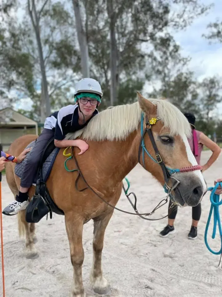 Person smiling on a tan horse with white mane, wearing a helmet. Outdoor setting.