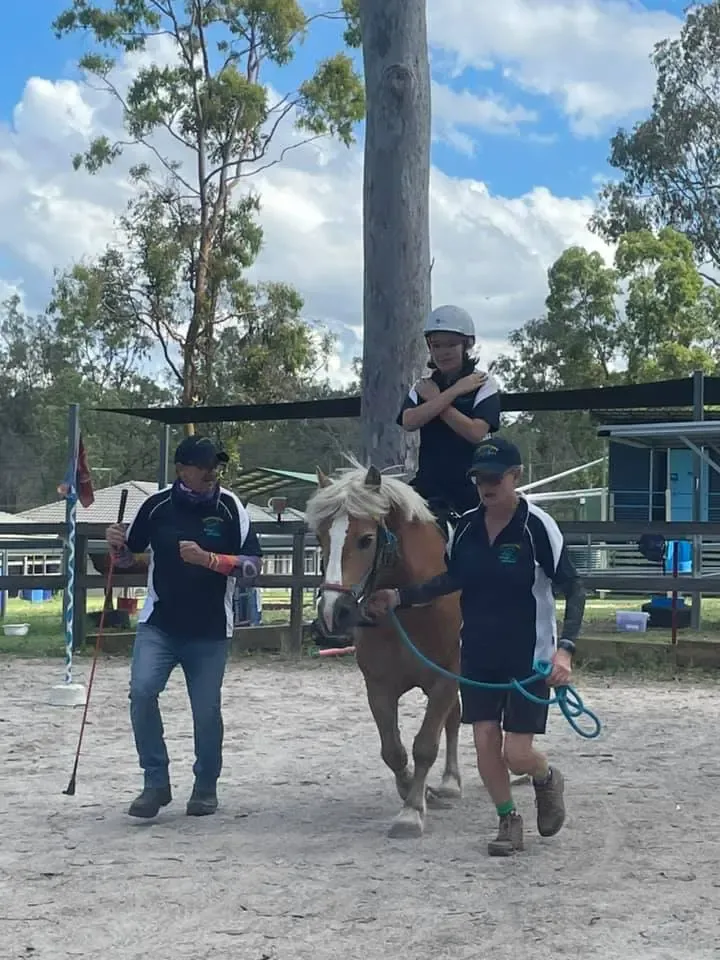 A person rides a pony led by two people in a riding arena on a sunny day.