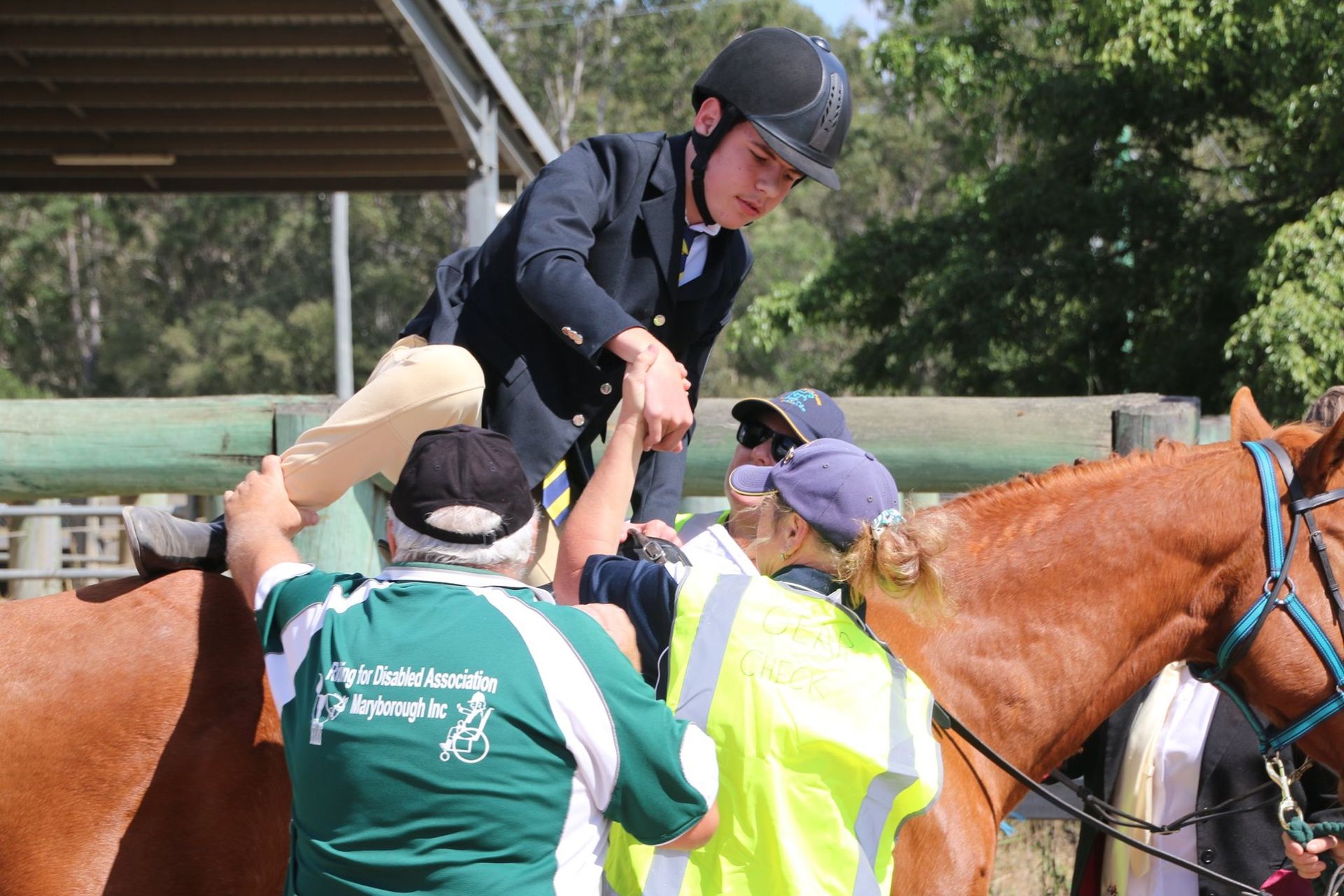 Rider being assisted onto a brown horse by a group of people at an outdoor equestrian event.
