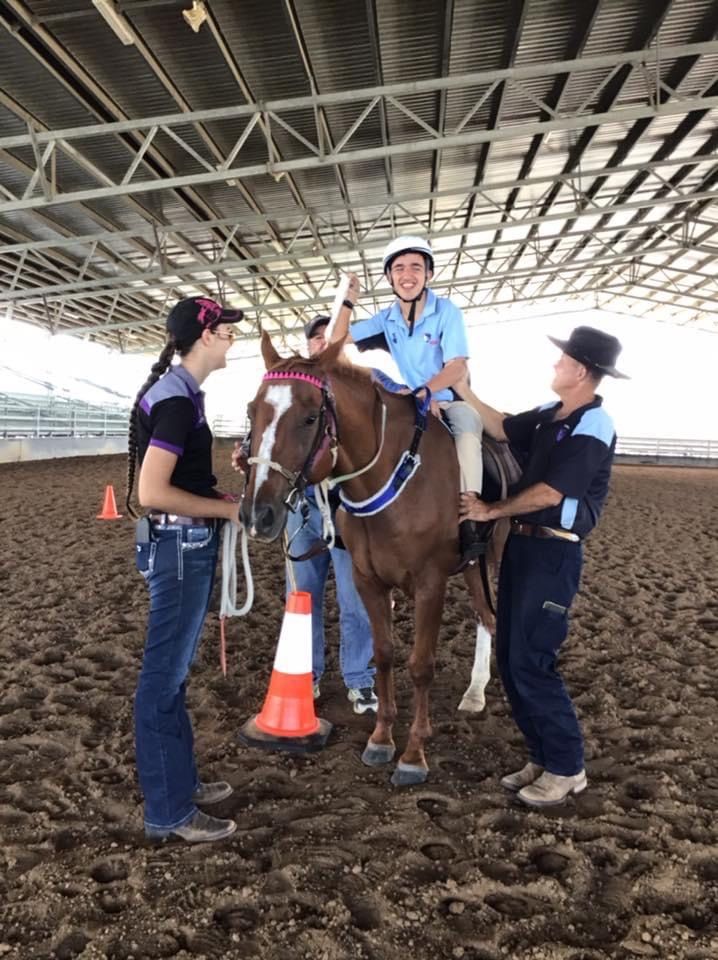 Person riding a horse in arena, assisted by two people. White hard hat, blue shirt on rider. Horse is brown with a pink bridle.