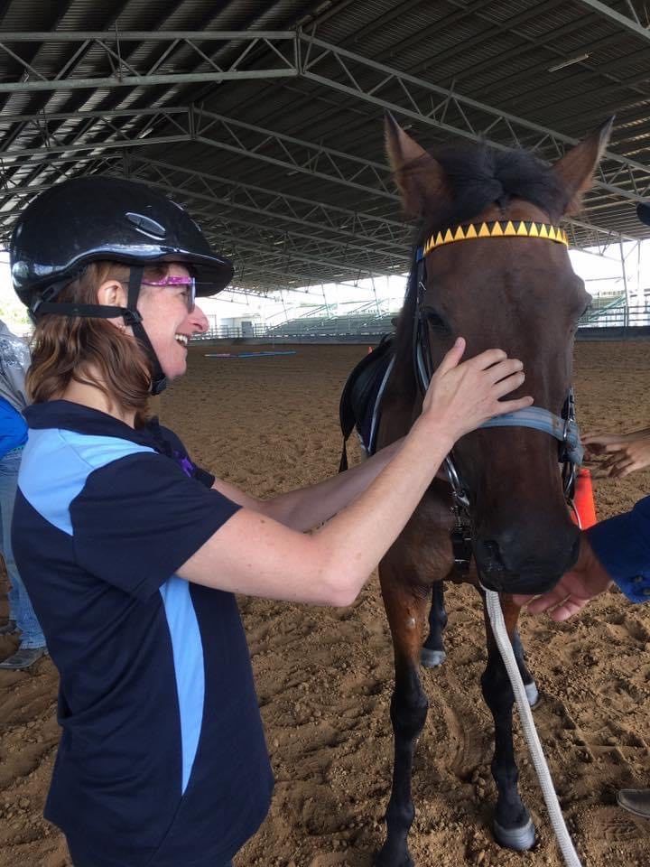 Woman in helmet pets a brown horse's face indoors; both appear happy.