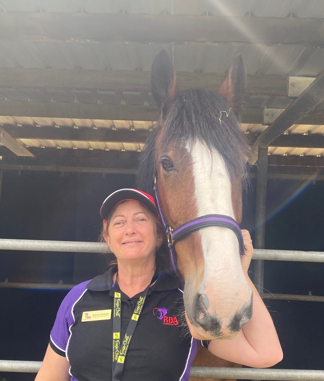 Woman in a purple-accented polo shirt and cap smiles next to a brown and white horse with a purple halter, in a stable.