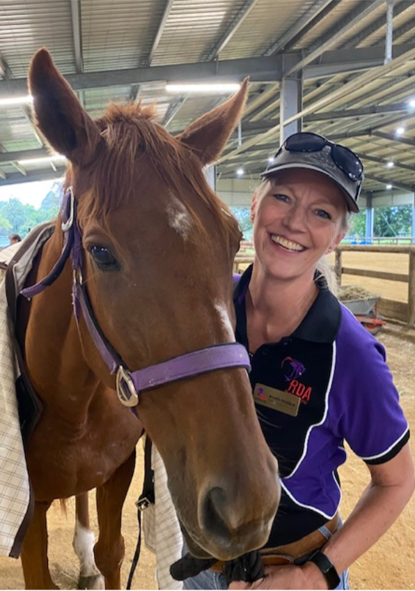 Woman smiles next to a brown horse in an indoor riding arena; woman wears a hat and polo shirt.
