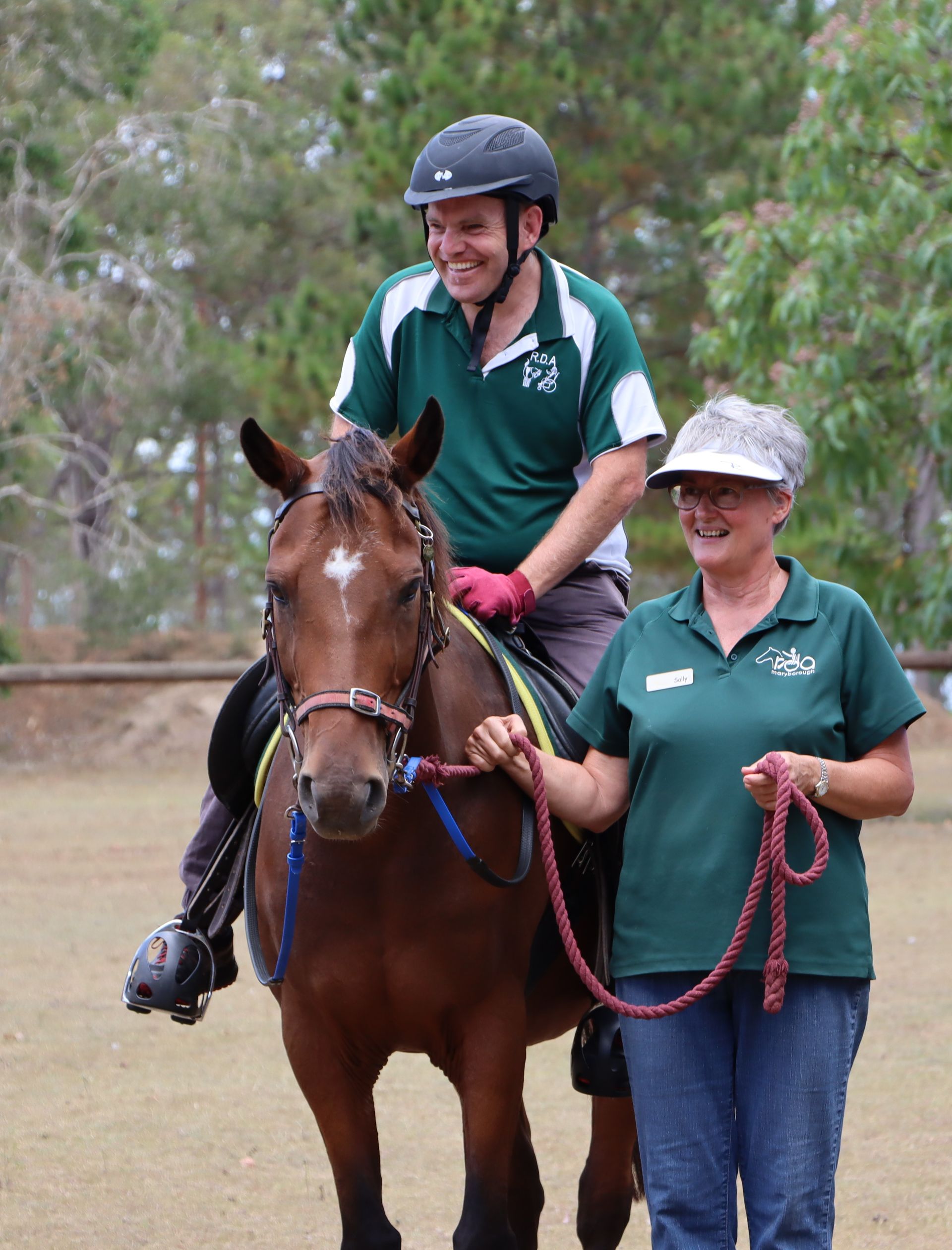 Man riding a brown horse guided by a woman; both wear green shirts. Outdoors.