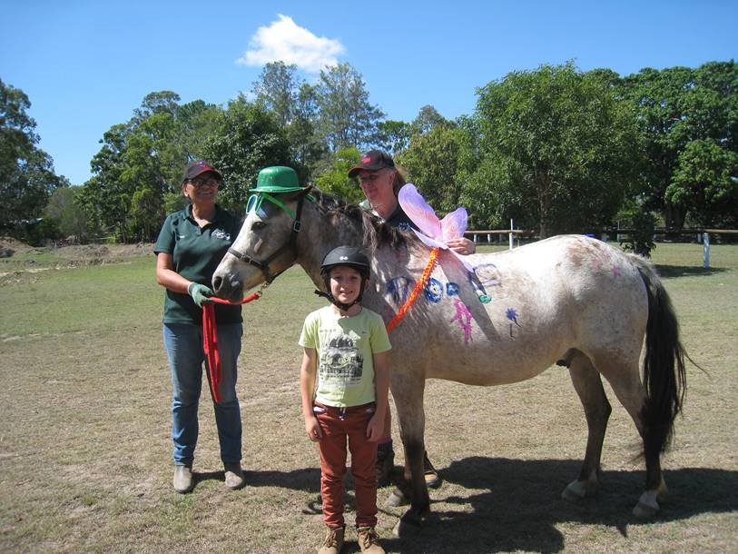 A person and child pose with a decorated horse outdoors; the horse has a green hat and wings.