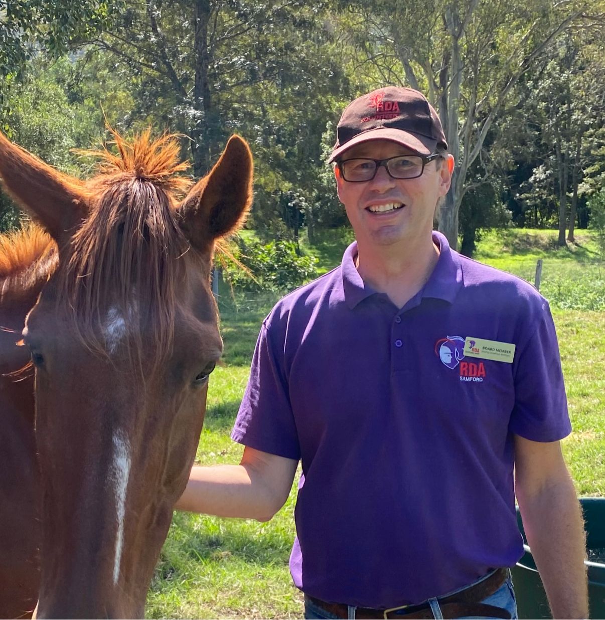 Man in purple shirt with glasses and horse in outdoor setting.